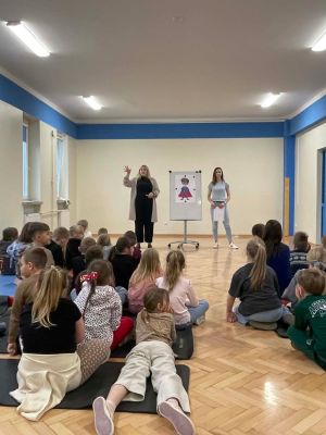 Uczniowie klas pierwszych aktywnie uczestniczą w warsztatach, siedząc na matach i słuchając prowadzących, kt&oacute;re wprowadzają ich w tematykę zajęć. 