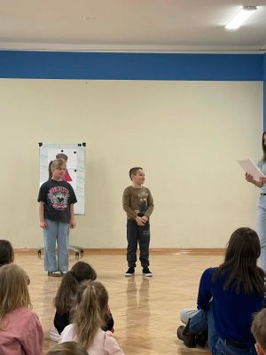 Na zdjęciu uczniowie biorą udział w scenkach, podczas kt&oacute;rych uczą się zasad prawidłowej komunikacji, a prowadząca wspiera ich i odczytuje przygotowane treści. 