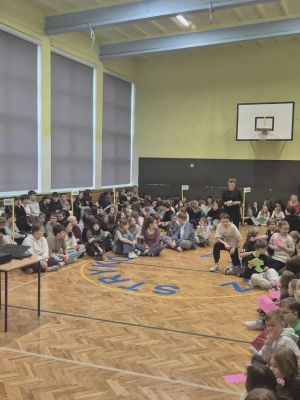 Uczniowie r&oacute;żnych klas zgromadzeni są w sali gimnastycznej, gdzie siedząc na podłodze uczestniczą w apelu szkolnym.